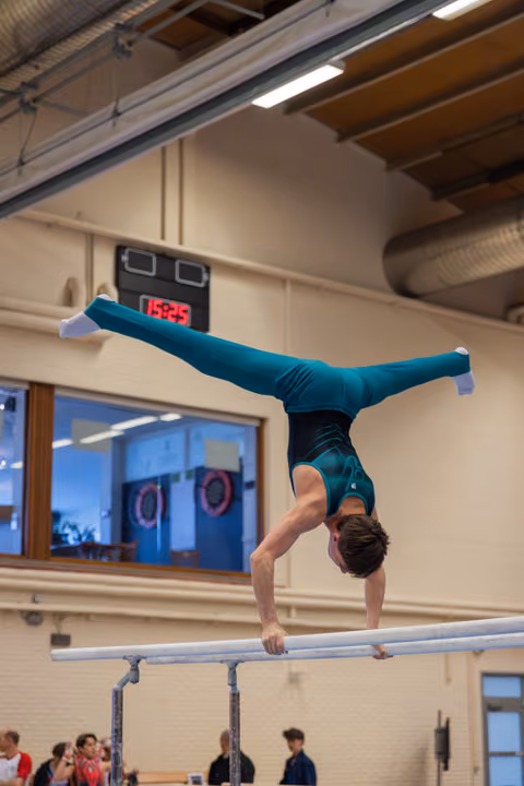 Male gymnast performs a handstand with legs split on parallel bars, wearing a teal leotard, in an indoor gymnasium.
