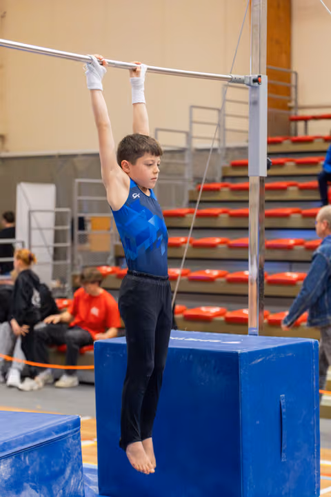 Focused young boy hangs from a horizontal bar in a blue leotard, feet lifted, competing in a gymnastics hall.