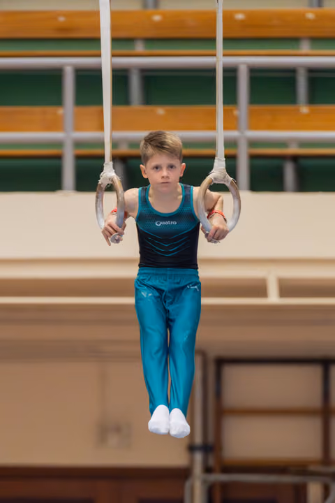 Focused young boy holds steady on gymnastics rings, wearing teal Quatro leotard, legs tucked, intense concentration on his face.