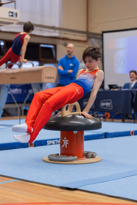 Young male gymnast performs focused pommel horse routine, legs extended, in a blue gymnasium with coach watching behind.
