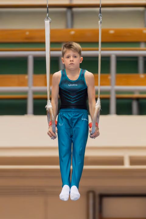 Young boy gymnast holds still rings with focused expression, wearing teal Quatro leotard in gymnasium