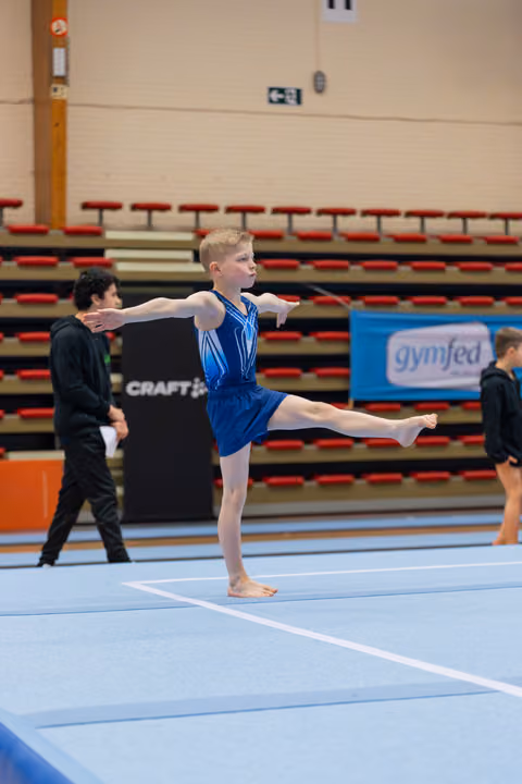 Young gymnast in blue leotard performs a balance pose on floor, arms outstretched, leg raised, focused expression.