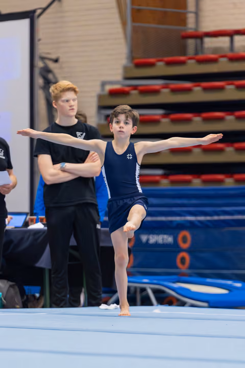 Young boy gymnast balances on one leg, arms outstretched, focused expression during floor exercise while coach watches behind.