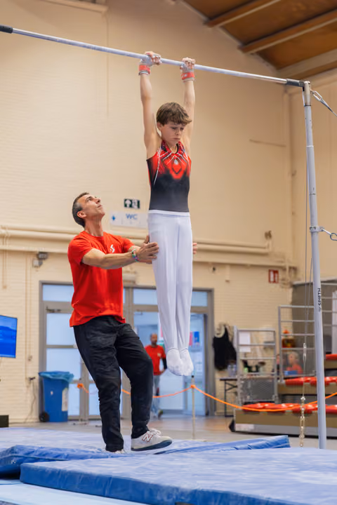 A coach steadies a young male gymnast hanging from the high bar, looking up attentively in an indoor gymnastics hall.