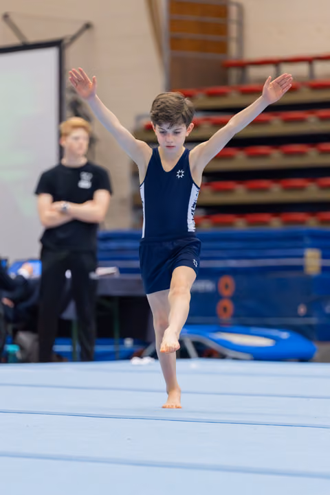 Young male gymnast performs floor routine on one foot, arms raised, with focused expression. Coach watches in background.
