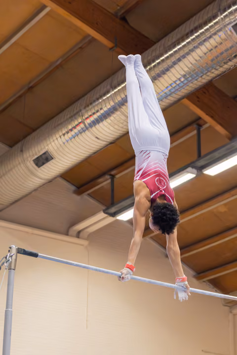 Male gymnast holds a perfect handstand on the high bar, body fully extended, in an indoor gymnasium.