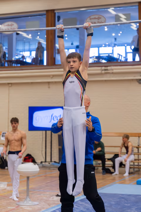Young male gymnast grips the high bar with intense focus while a coach in blue steadies him from below in a gymnasium.