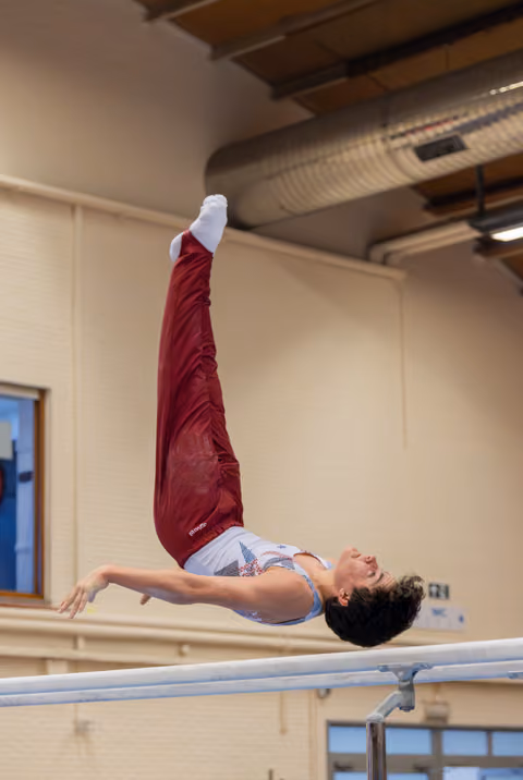Young gymnast performs a dramatic back arch release on parallel bars, legs extended upward, expression focused and intense.