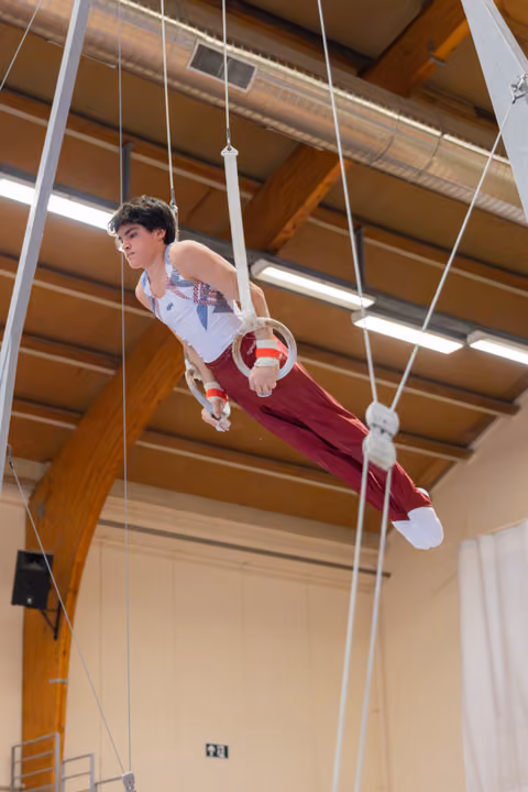 Young male gymnast performs on still rings, body horizontal, focused expression in a wooden-beamed gymnasium.
