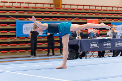 Young gymnast performs a precise arabesque on the floor mat, arms raised gracefully, judges scoring at the table behind.