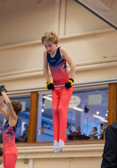 Young boy in red and navy leotard grips the horizontal bar with intense focus during a gymnastics event.