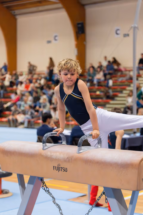 Determined young gymnast grips pommel horse handles mid-routine, legs extended, concentration visible on his face. Indoor arena with spectators.