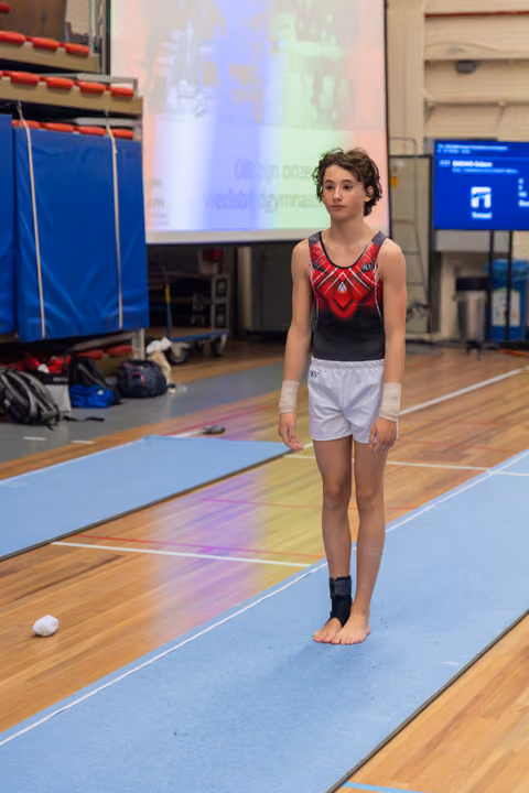 Young gymnast stands composed on the floor mat, wearing a red leotard and wrist wraps, preparing for a routine.