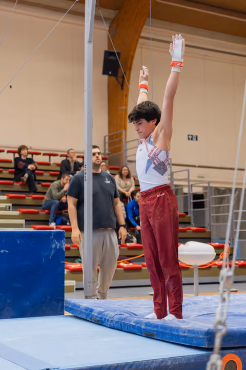 Young male gymnast stands tall on mat, arms raised holding rings, focused expression, coach watching in background.