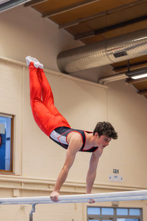 Young male gymnast performs a dramatic horizontal hold on parallel bars, body parallel to the floor, legs raised high.