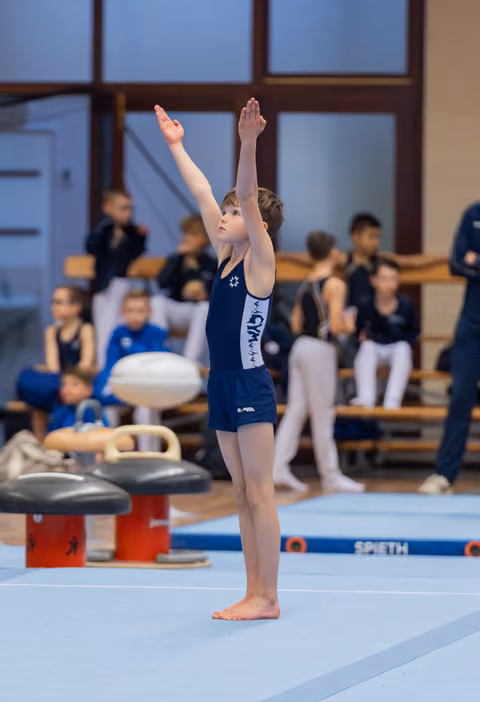 Young gymnast stands tall on the mat, arms raised in a proud salute, facing the judges in an indoor gymnastics hall.