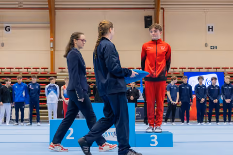 Young gymnast in red tracksuit stands on 3rd place podium as officials approach to present award in sports hall.