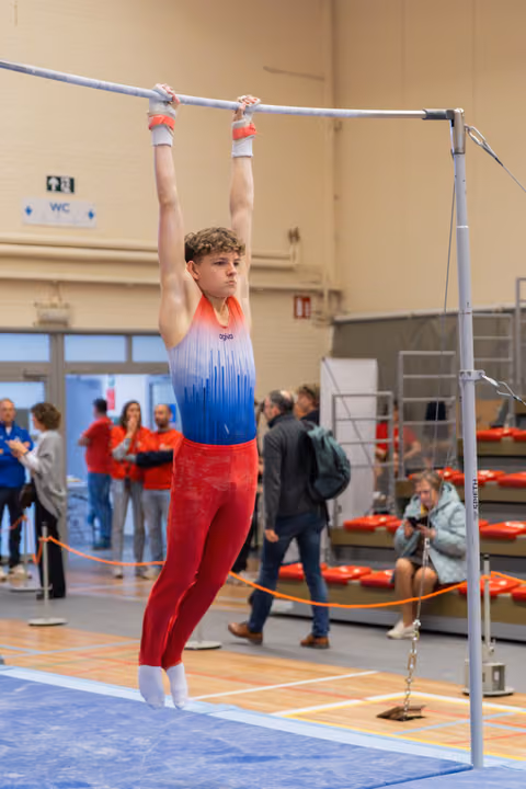 Focused teenage male gymnast hangs on the high bar with intense concentration, wearing a red and blue leotard indoors.
