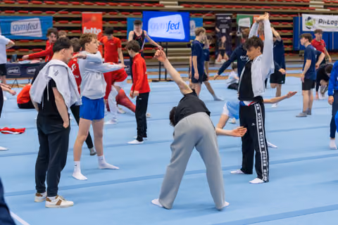 Young male gymnasts warm up on blue floor mats, stretching and reaching upward in a busy competition hall with Gymfed banners.