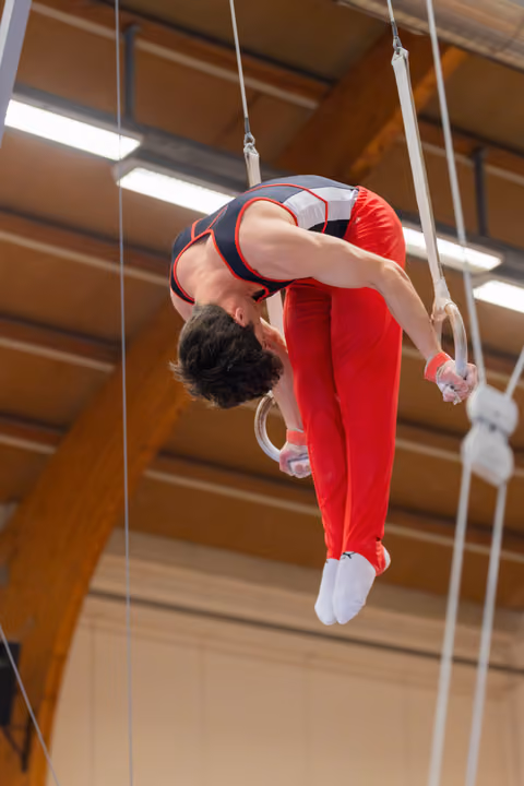 Male gymnast in red and black leotard performs a dramatic backbend on still rings in an indoor gymnasium.