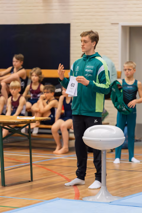 Young male gymnast wearing number 63 stands confidently, gesturing while holding a score card in a gymnasium.