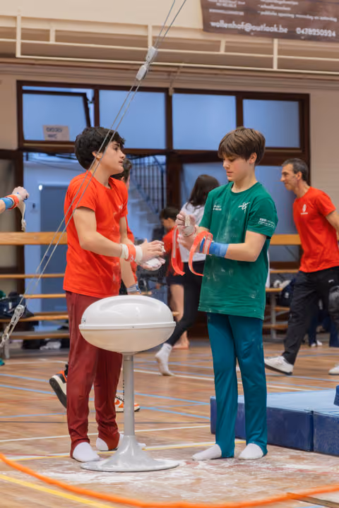 Two young gymnasts apply chalk before their event, the boy in green holding red rings while his teammate assists, both focused.