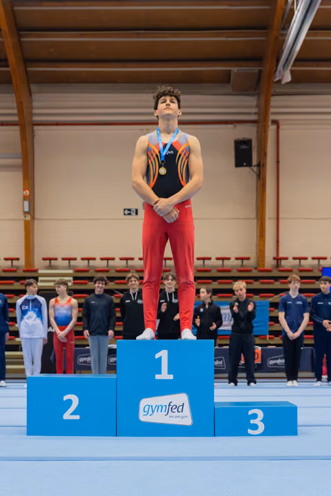 Young male gymnast stands tall on the 1st place podium, chin raised, gold medal around his neck at a Gymfed event.