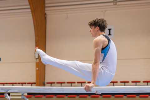 Young male gymnast performs a straddle L-sit on parallel bars, focused and controlled, in an indoor gymnasium.