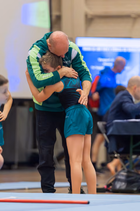 Bald coach in green jacket embraces young gymnast in teal leotard on floor mat, a moment of comfort or celebration.