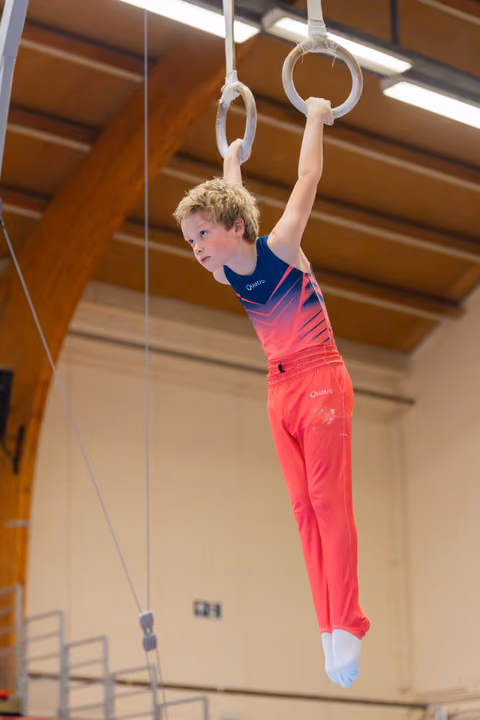 Young boy in red and blue Quattro kit hangs with intense focus on gymnastic rings in a sports hall.