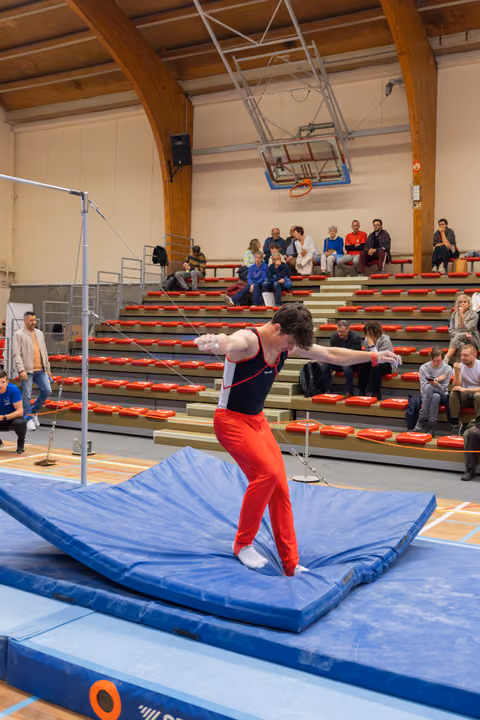 Young male gymnast lands on blue mat, arms wide for balance, leaning forward with intense focus. Spectators watch from bleachers.