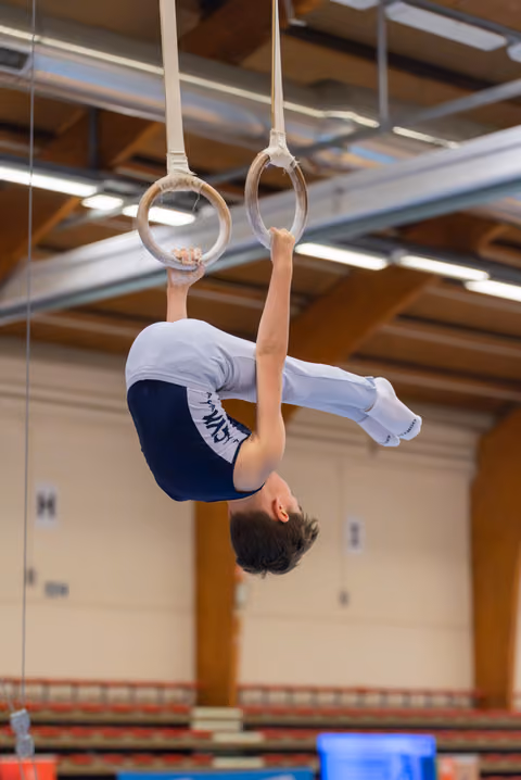 Young male gymnast performs an inverted hang on still rings, body curved with concentration, inside a sports hall.