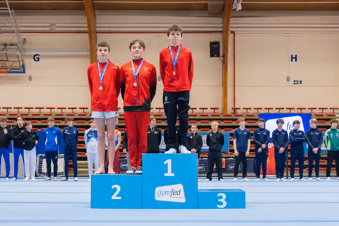 Three young male gymnasts stand proudly on a blue Gymfed podium, wearing red jackets and blue medals around their necks.