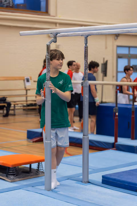 Young male gymnast in green shirt grips parallel bars, looking intently sideways in a busy gymnastics hall.