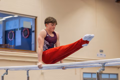 Young male gymnast performs a straddle hold on parallel bars, legs raised high, expression focused and intense indoors.