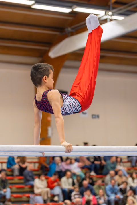 Young male gymnast holds an inverted pike position on parallel bars, legs raised high in red trousers, focused expression.