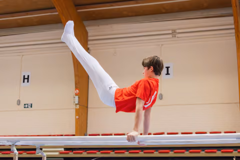 Young boy in red shirt performs a leg raise on parallel bars, showing intense focus and strong body control in a gymnasium.