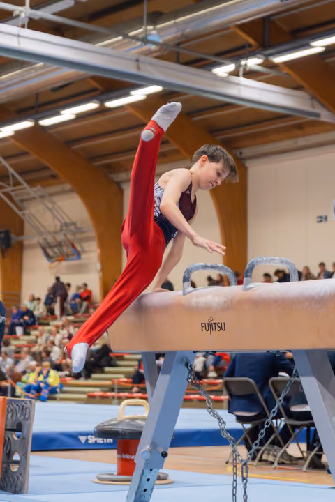Young male gymnast performs a powerful scissor move on the pommel horse, leg extended high, intense focus on his face.