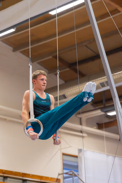 Young male gymnast performs a leg raise on still rings, focused expression, wearing teal leotard in an indoor gymnasium.