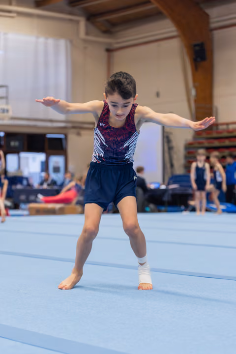 Young male gymnast performs floor routine, arms outstretched for balance, focused expression, ankle bandaged, in a gymnasium.