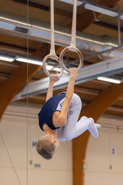 Young male gymnast hangs inverted on still rings, body tucked, showing intense focus and strength in a gymnasium.