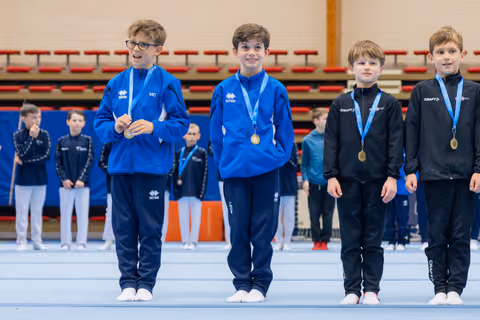 Four young gymnasts stand proudly on the podium, wearing gold and silver medals, smiling in blue and black tracksuits.