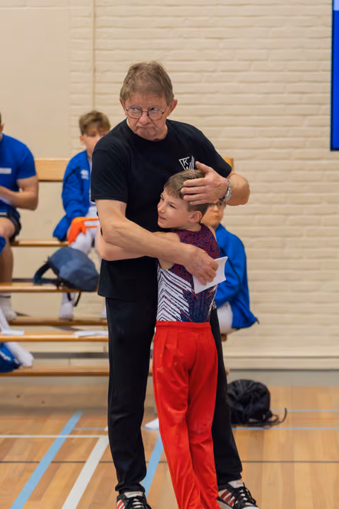 An older coach in black embraces a young gymnast in red pants, offering reassurance in a sports hall.