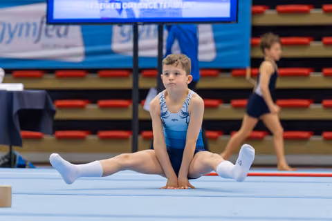 Young boy in blue leotard holds a wide split on the floor mat, looking up with calm concentration during a gymnastics event.
