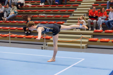 Young gymnast holds a precise arabesque balance on the floor mat, arms forward, leg raised, focused expression.