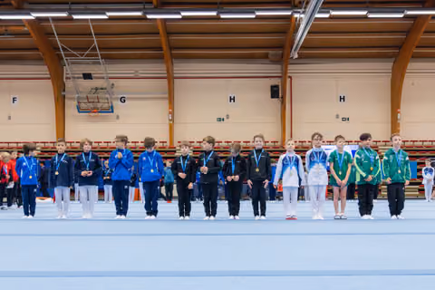 Young gymnasts stand proudly in a line wearing medals at an indoor gymnastics competition, dressed in colorful team tracksuits.
