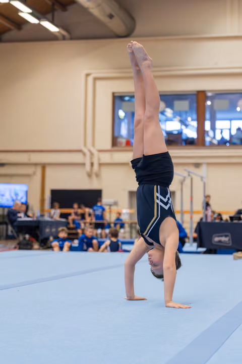 Shirtless boy performs a focused handstand on the blue floor mat during a gymnastics competition.