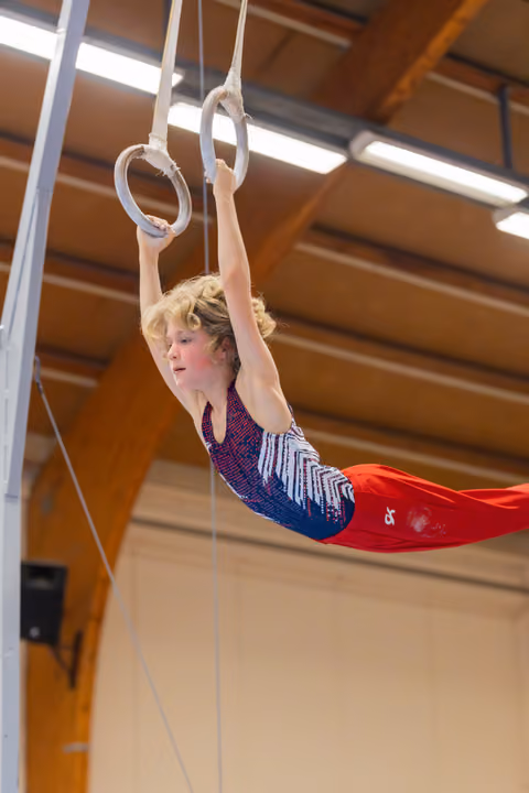 Young gymnast holds a horizontal position on still rings, focused expression, in a wood-beamed sports hall.