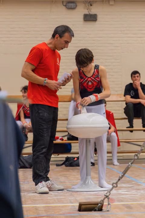 A focused coach in red helps a young gymnast apply chalk to his wrists before a pommel horse routine in a gymnasium.