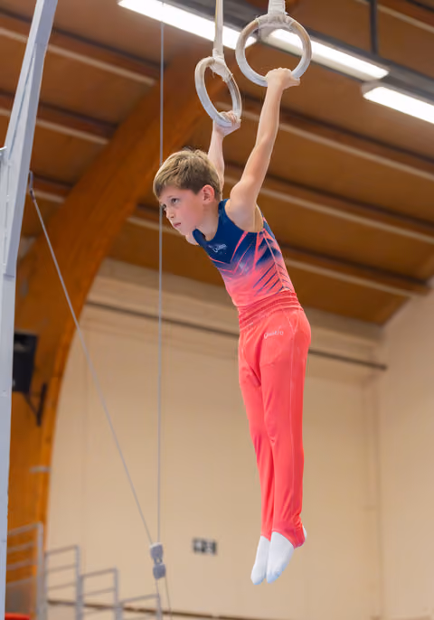 Focused young gymnast hangs from still rings in a sports hall, wearing a blue and pink leotard with red trousers.